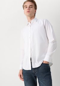 Camisa blanca de manga larga con botones y cuello clásico, textura suave y un discreto detalle de logo en el pecho. Combinada con jeans de mezclilla oscura.