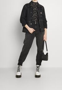 Schwarze Jeansjacke mit weißen Nähten, schwarzes Oberteil mit floralem Muster, schwarze Jeans, weiße Schnürstiefel mit klobigen Sohlen und eine kleine schwarze Handtasche.