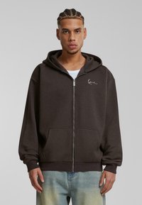 Sudadera con cremallera de color marrón, con capucha ajustable, bolsillos delanteros y logo bordado en el pecho. Tejido suave con un corte holgado y puños acanalados.