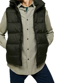 Mörkgrå pufferväst med hög krage, dragkedja fram och fyrkantiga lappar. Bärs över en ljusfärgad skjorta med knappar.