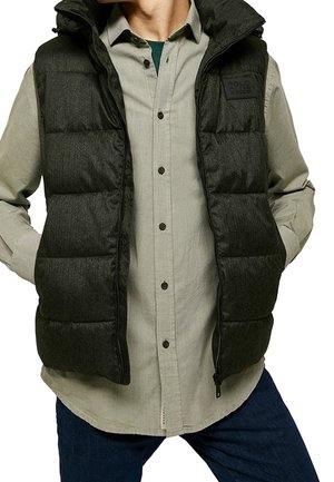 Chaleco acolchado gris oscuro con un alto cuello, cierre frontal con cremallera y parches cuadrados. Llevado sobre una camisa de botones de color claro.