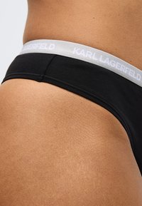 Čierne elastické bavlnené bikiny so sivým elastickým pásom s nápisom "KARL LAGERFELD" bielou farbou. Detailný záber zdôrazňuje textúru a strih.