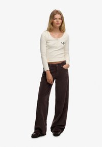 Camisa henley de manga larga color crema con botones y logo, combinada con pantalones de pana marrón oscuro de pierna ancha y zapatos negros, diseño minimalista.