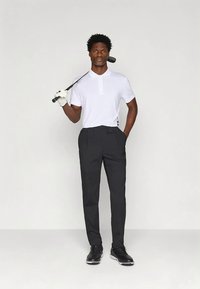 Homem com luva de golfe branca, camisa polo branca, calças pretas e sapatos de golfe pretos segura um taco de golfe sobre o ombro em fundo simples.