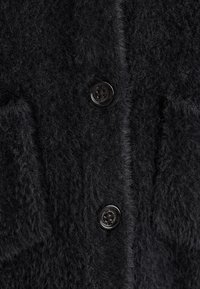 Schwarze, flauschige Strickjacke mit zwei großen Taschen und glänzenden schwarzen Knöpfen, die eine weiche Textur und eine lockere, entspannte Passform zeigt.