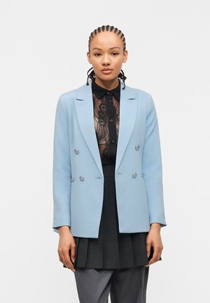Femme aux cheveux tressés portant un blazer croisé bleu clair sur une chemise noire en dentelle transparente et une jupe plissée avec pantalon gris, debout droite.