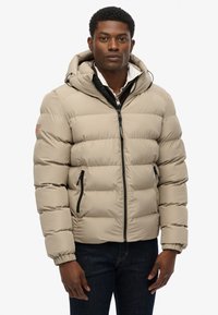 Beige gewatteerde jas met capuchon, voorzien van een ritssluiting aan de voorkant, ritszakken en elastische manchetten. Zachte textuur, gewatteerd ontwerp en branding op de mouw.