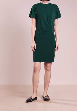 Vrouw draagt een donkergroene jurk met korte mouwen en een lengte tot aan de knie, met zwarte, gestikte puntige platte schoenen, staand tegen een effen beige achtergrond.