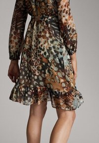 Femme portant une robe transparente à motif animalier, manches longues et ourlet à volants, vue de dos jusqu'aux genoux.