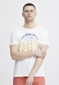 Weißes Baumwoll-T-Shirt mit kreisförmigem Grafiken in blau und gelber Schrift, das Phrasen wie "Hier kommt die Sonne" zeigt. Modell mit kurzen Haaren und verschränkten Armen.