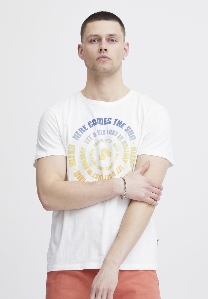 Weißes Baumwoll-T-Shirt mit kreisförmigem Grafiken in blau und gelber Schrift, das Phrasen wie "Hier kommt die Sonne" zeigt. Modell mit kurzen Haaren und verschränkten Armen.