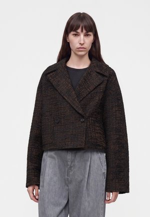 Femme aux cheveux bruns raides porte un manteau court texturé foncé par-dessus un haut noir et un pantalon gris ample sur un fond uni.