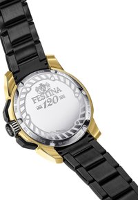 Črna kovinska ura z zlatimi detajli, okrogla ohišje in strukturiran pas. Na hrbtni strani je srebrna površina z vtisnjenim logotipom in podatki o vodoodpornosti.