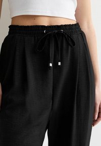 Schwarze elastische Hose mit Gummizug und silbernen Metallenden. Der Stoff scheint weich mit einer leichten Textur und hat eine lockere Passform.