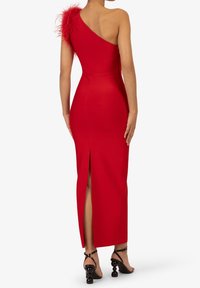 Robe rouge à une épaule avec un accent en plumes, silhouette ajustée, fente sur le côté et tissu lisse. Portée avec des sandales à talons carrés noires.