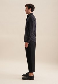 Camicia nera su misura, vestibilità rilassata, maniche lunghe; abbinata a pantaloni neri a vita alta. Modello che indossa scarpe nere, in posa di profilo. Sfondo neutro.