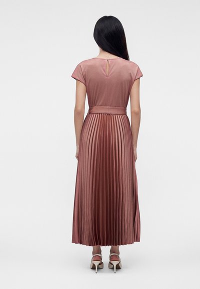 Femme aux longs cheveux foncés portant une robe midi plissée rose poussière avec des manches courtes et des sandales blanches à talons hauts, debout, de dos.