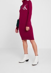 Fuchsia stickad klänning med hög krage och långa ärmar, med en vit "CK"-logotyp. Bärs med vita klackade ankelskor.