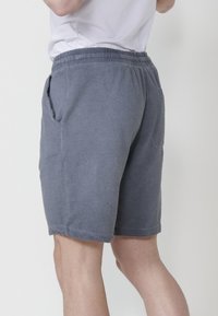 Man som bär texturerade grå shorts och vit skjorta, visad från midjan neråt med ena handen nära ansiktet, mot en enhetlig bakgrund.