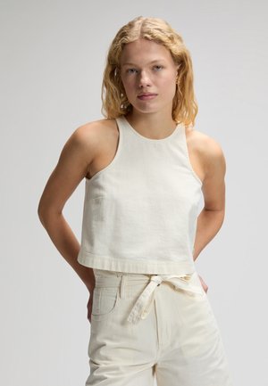 Blondynka ubrana w białą, bezrękawnikową krótka bluzkę i białe spodnie z wysokim stanem związane kokardą, stojąca z rękami za plecami.