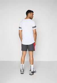 T-shirt blanche à manches courtes avec des rayures noires sur les manches, associée à un short gris et rouge, des chaussettes blanches et des chaussures de sport grises.
