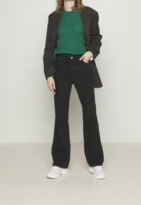 Persona che indossa un maglione di maglia verde, una giacca scura, pantaloni neri a vita alta e sneakers bianche, in piedi contro uno sfondo neutro.