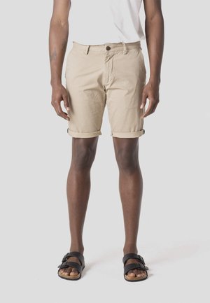 Férfi bézs színű, felhajtott rövidnadrágban, fehér pólóban és fekete papucsban, egy egyszerű világos háttér előtt áll.