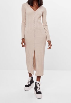 Femme portant une robe midi beige côtelée boutonnée à manches longues, associée à des baskets compensées noires et blanches à lacets.