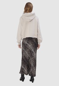 Vrouw draagt een beige cropped hoodie en een lange luipaardprint rok met zwarte laarzen met hakken, staand met haar rug naar de camera.