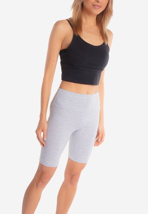 Kobieta ubrana w czarny krótki top bez rękawów i jasnoszare, wysokie spodenki rowerowe, stojąca na białym tle.