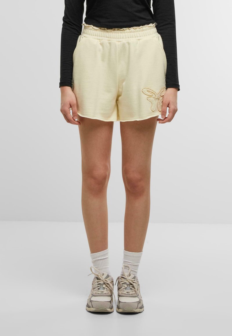 Shorts couleur crème avec une taille élastique, des poches latérales et un détail brodé de papillon décoratif sur l'ourlet droit.