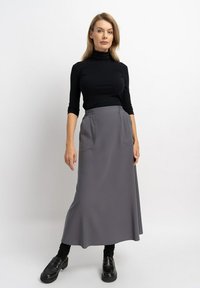 Grauer, knie-langer Rock mit zwei vorderen Taschen, elastischem Bund, kombiniert mit einem figurbetonten schwarzen Rollkragenpullover und schwarzen Schuhen. Einfarbiger Hintergrund.