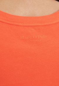 Īsām piedurknēm, spilgti oranža kokvilnas krekls ar ribotu apkakli. Uz gludas virsmas tuvumā apkaklei ir ieplākots teksts "Marc O'Polo".