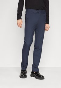 Pantalones azul marino con un diseño de frente plano, ajuste slim y trabillas para cinturón, combinados con mocasines negros brillantes y una camisa negra de manga larga.