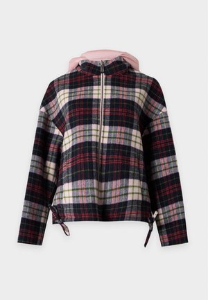 Femme portant une veste à carreaux noire, blanche, rouge et verte avec une capuche rose, un jean large bleu clair et des chaussures beiges, debout sur un fond uni.