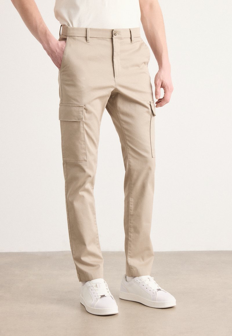 Beige cargobyxor med sidofickor, figursydd design, bomullsmaterial, raka ben, i kombination med vita sneakers på en neutral bakgrund.