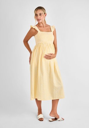 Mujer embarazada de pie, vestida con un vestido midi sin mangas de cuadros amarillos y sandalias blancas sin cierre, con una mano en su barriga.