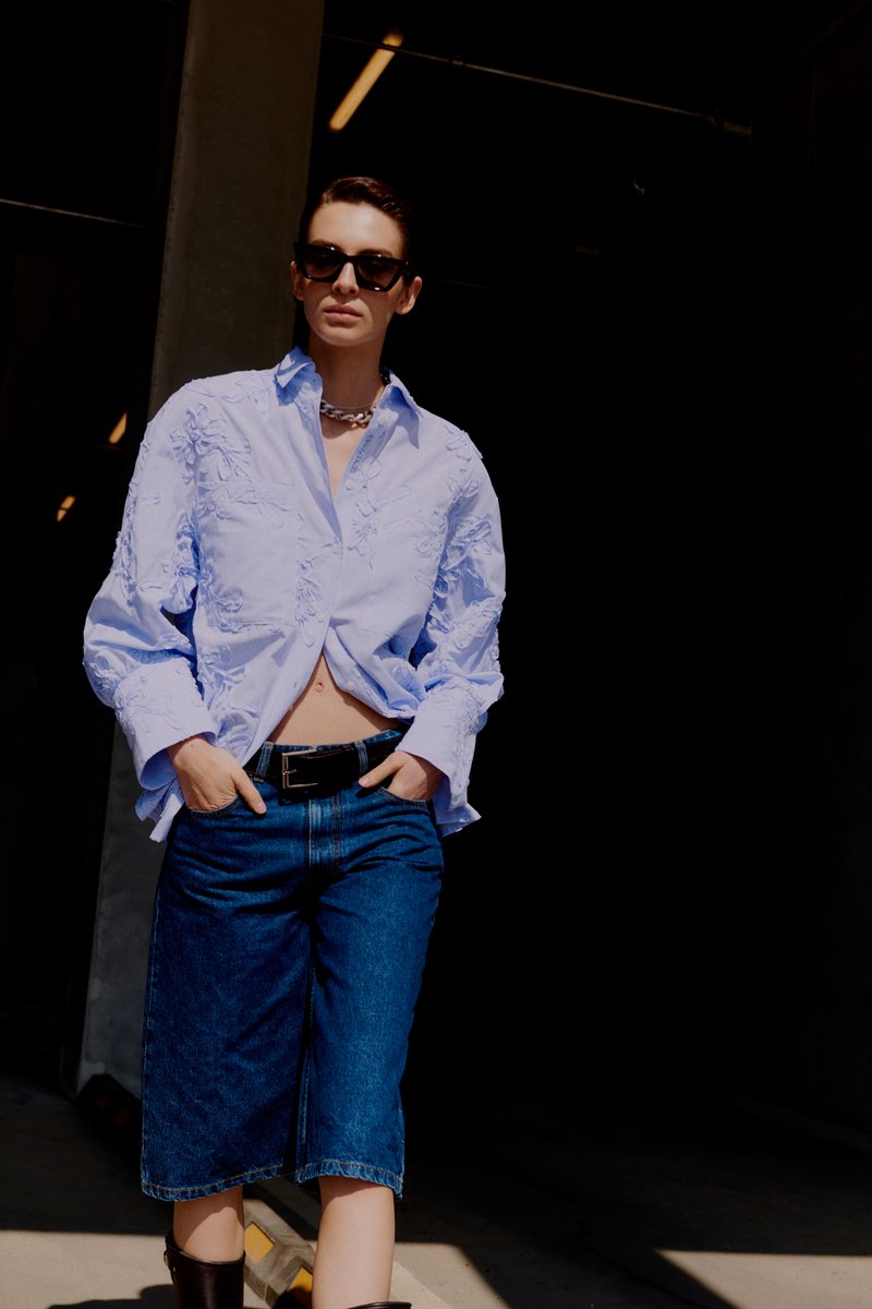 Γυναίκα που φοράει ανοιχτό μπλε κεντημένο πουκάμισο, σκούρα μπλε τζιν σορτς, μαύρη ζώνη, γυαλιά ηλίου και κολιέ με αλυσίδα, στέκεται στη σκιά.