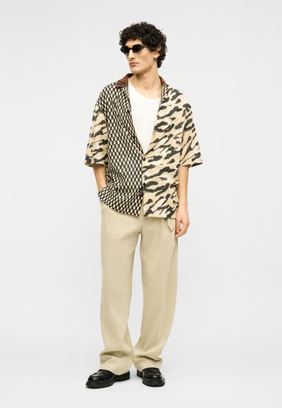 Άνδρας που φοράει πουκάμισο με ανάμεικτα σχέδια, μπεζ παντελόνι, μαύρα παπούτσια, μικρά μαύρα γυαλιά ηλίου, στέκεται με το ένα χέρι στην τσέπη μπροστά σε λευκό φόντο.