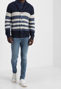 Strickjacke mit navyblauen und cremefarbenen horizontalen Streifen, Schal-Kragen, Knopfleiste, über Jeans getragen, kombiniert mit weißen Sneakern.