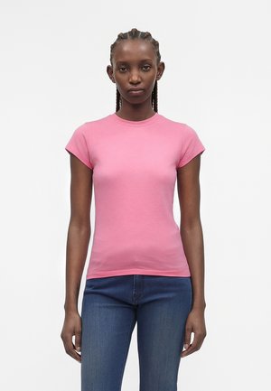 Kvinde med flettet hår iført en tætsiddende kortærmet pink t-shirt og blå jeans, stående mod en ensfarvet hvid baggrund.