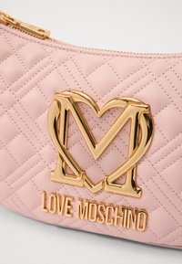 Kvadričasta ružičasta sintetička kožna torba s zlatnim metalnim logotipom u obliku srca i urezanim tekstom "LOVE MOSCHINO" na prednjoj strani.