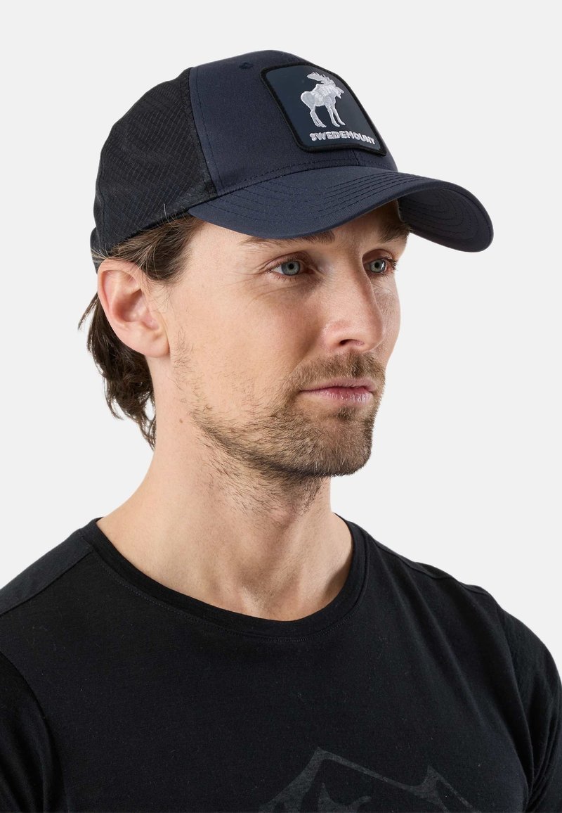 Homme avec une légère barbe portant une casquette noire ornée d'un logo élan blanc et du mot « SwedenMount », ainsi qu'un t-shirt noir à col rond.