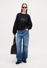 Sweatshirt corto nero con applicazioni di strass, jeans blu a gamba larga, borsa nera e scarpe nere. La modella indossa occhiali da sole scuri.