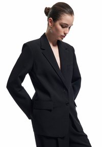Vrouw met een bun en een oversized zwarte blazer, draagt gouden ringoorbellen en kijkt naar beneden tegen een plain witte achtergrond.