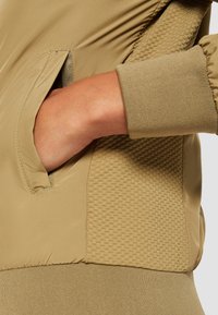 Main dans la poche d'une veste kaki montrant un poignet côtelé, un panneau latéral texturé et un tissu lisse sur les zones de la poche et de la ceinture.