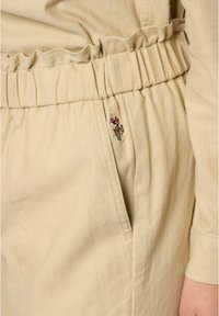 Beige, lichtgewicht stof met een gerimpelde tailleband en een rimpeldetail. Heeft een decoratieve geborduurde accent op de zak.