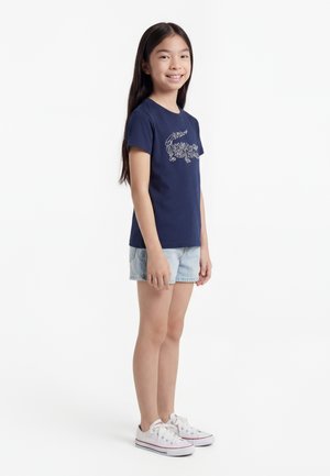 Junges Mädchen steht lächelnd da, trägt ein dunkelblaues T-Shirt mit Grafik, hellblaue Jeans-Shorts und weiße Turnschuhe vor weißem Hintergrund.
