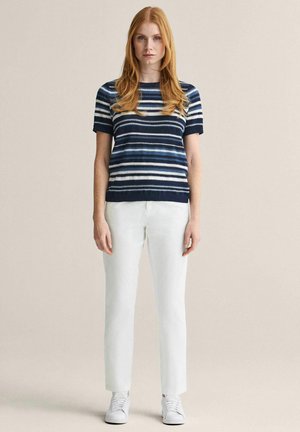 Nainen seisoo pukeutuneena tummansiniseen raidalliseen lyhythihaiseen villapaitaan, valkoisiin housuihin ja valkoisiin tennareihin yksinkertaisen beigen taustan edessä.