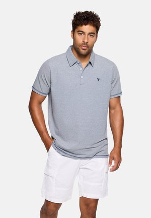 Hombre con cabello corto y rizado, vistiendo una camiseta polo a rayas azul claro y pantalones cortos tipo cargo blancos hasta la rodilla, con las manos en los bolsillos, fondo neutro.